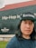 A person is standing outdoors wearing a black cap with 'Racionais' written on it. Behind them is a large brick wall painted with text, 'Hip-Hop is...' and 'hulu RapCaviar Presents'. The wall is divided into two sections with black and red paint. There's a small patch of green grass in the foreground.