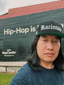 A person is standing outdoors wearing a black cap with 'Racionais' written on it. Behind them is a large brick wall painted with text, 'Hip-Hop is...' and 'hulu RapCaviar Presents'. The wall is divided into two sections with black and red paint. There's a small patch of green grass in the foreground.