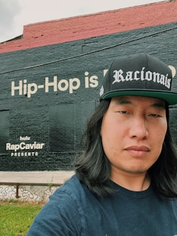 A person is standing outdoors wearing a black cap with 'Racionais' written on it. Behind them is a large brick wall painted with text, 'Hip-Hop is...' and 'hulu RapCaviar Presents'. The wall is divided into two sections with black and red paint. There's a small patch of green grass in the foreground.
