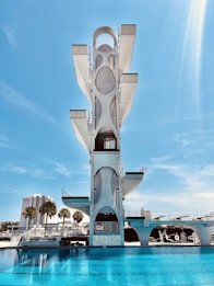 A tall, modern diving platform structure stands against a clear blue sky. The platform has multiple levels with geometric cutouts and sleek design elements. Below it is a large, inviting swimming pool with clear blue water. In the background, there are palm trees and a few multi-story buildings, adding an urban backdrop to the scene.