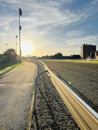 A race track bordered by a metal railing with a paved path alongside and grassy areas. The sun is either setting or rising, casting a warm golden light on the scene. There are tall floodlights along the path and buildings in the background. Additionally, trees and a partly cloudy sky enhance the atmosphere.