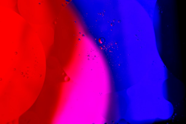 Colorful abstract pattern with vibrant red, pink, and blue hues blending together. Tiny bubbles or droplets are scattered across the surface, adding texture and depth.