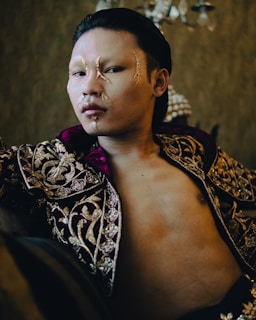 A person with long, slicked-back hair wears an elaborate, ornate garment with intricate gold embroidery on deep purple fabric. The face has artistic makeup featuring golden accents and a shiny, smooth complexion. The background is a lavish, dimly lit room with a luxurious ambiance, adding an air of elegance and mystery.