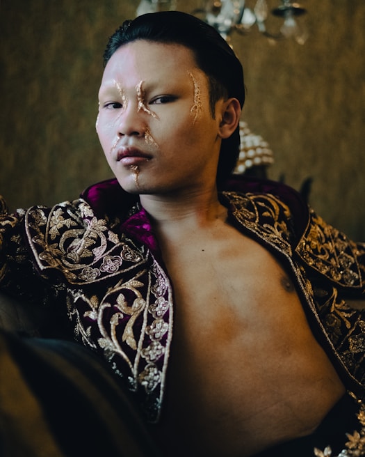 A person with long, slicked-back hair wears an elaborate, ornate garment with intricate gold embroidery on deep purple fabric. The face has artistic makeup featuring golden accents and a shiny, smooth complexion. The background is a lavish, dimly lit room with a luxurious ambiance, adding an air of elegance and mystery.