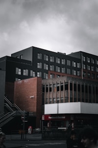 A multi-story urban building with a modern design features a combination of dark panels and brickwork. The lower section houses stores labeled 'Furniture & Electrical.' Overcast skies add a somber atmosphere, with muted natural light highlighting the architectural details.