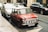 A classic red Mini Cooper is parked on the side of a city street. It features several round headlights and a chrome bumper. Other cars are parked nearby, and the street is lined with buildings.