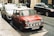 A classic red Mini Cooper is parked on the side of a city street. It features several round headlights and a chrome bumper. Other cars are parked nearby, and the street is lined with buildings.