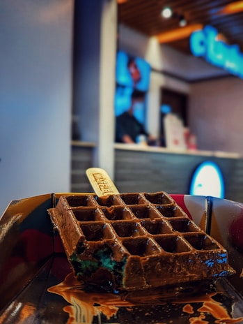 A chocolate waffle on a stick is displayed on a dark-colored tray with melting chocolate drips. The background is blurred, showing part of a modern interior with shelves and a person at a counter.