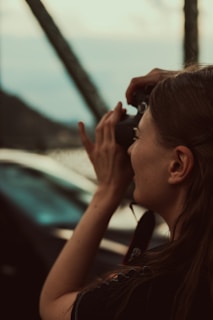 A person with long hair holds a camera up to their face, seemingly taking a photograph. The setting appears outdoors, with a blurred background indicating possibly a mountainous or countryside landscape. The lighting is warm, suggesting late afternoon or early evening.