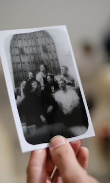 A hand is holding a black and white photograph featuring a group of people posing in front of a large, ornate wooden door. The group appears to be smiling and interacting joyfully with each other.