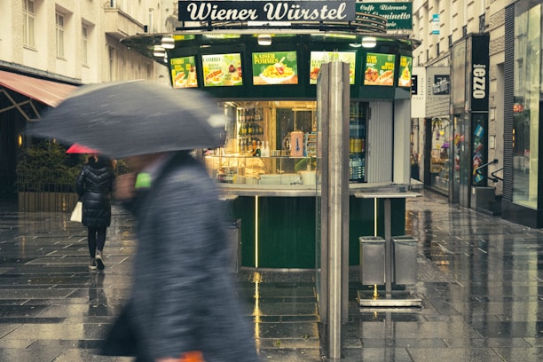 A street scene featuring a food kiosk with the sign 'Wiener Würstel' surrounded by wet pavement, suggesting recent rain. People are walking by, some holding umbrellas. The kiosk displays food advertisements, and the setting appears to be an urban shopping area with stores on both sides.