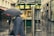 A street scene featuring a food kiosk with the sign 'Wiener Würstel' surrounded by wet pavement, suggesting recent rain. People are walking by, some holding umbrellas. The kiosk displays food advertisements, and the setting appears to be an urban shopping area with stores on both sides.