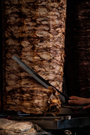 A large vertical spit of layered, marinated meat is being sliced with a long knife. The meat has a golden-brown, cooked appearance, and there is a visible hand holding the knife. The scene indicates a typical method of preparing shawarma or gyro.