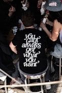 A group of people sits closely together, eating food from wrappers. One individual is prominently featured wearing a black t-shirt with white lettering that reads 'you can't always get what you want'. The surrounding area appears casual and informal, with stools and some takeaway food on a table.