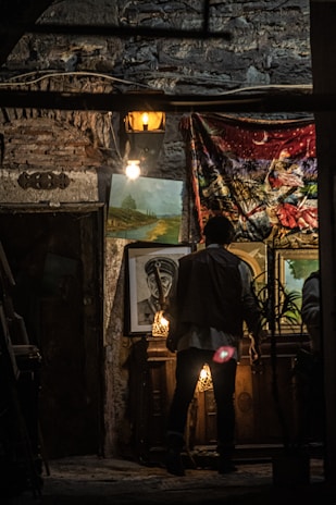 A dimly lit room displays various paintings against a textured stone wall. An individual stands in front of the artwork, examining it closely. Two light bulbs hang, adding a warm glow to the scene. A tapestry with vibrant colors partially covers the wall.