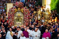 A large group of people gathers for a religious procession. At the center, there is an ornate structure adorned with flowers, possibly a religious reliquary or shrine, surrounded by clergy in ceremonial garments. The crowd includes people of all ages, intently participating in the event.