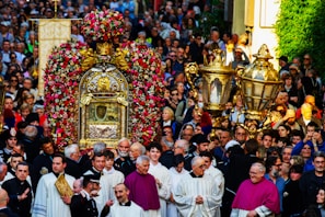 A large group of people gathers for a religious procession. At the center, there is an ornate structure adorned with flowers, possibly a religious reliquary or shrine, surrounded by clergy in ceremonial garments. The crowd includes people of all ages, intently participating in the event.