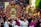 A large group of people gathers for a religious procession. At the center, there is an ornate structure adorned with flowers, possibly a religious reliquary or shrine, surrounded by clergy in ceremonial garments. The crowd includes people of all ages, intently participating in the event.