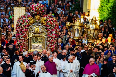A large group of people gathers for a religious procession. At the center, there is an ornate structure adorned with flowers, possibly a religious reliquary or shrine, surrounded by clergy in ceremonial garments. The crowd includes people of all ages, intently participating in the event.