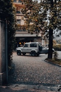 A rugged SUV is parked on a cobblestone driveway in front of a hotel with a classic European facade. Lush greenery, including trees with dense leaves, frames the scene, and soft, diffused lighting suggests an overcast day.