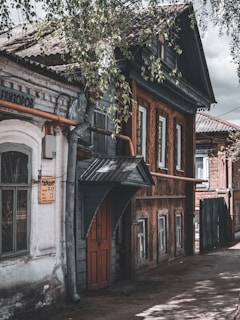 A rustic street scene featuring an old wooden house with intricate woodwork patterns on its exterior walls. The building has a sloped roof and a prominent overhang above a wooden door. Next to it stands a white brick structure with a sign on the wall. Leaves from overhanging trees partially obscure the view, adding a natural frame.