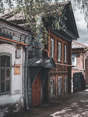 A rustic street scene featuring an old wooden house with intricate woodwork patterns on its exterior walls. The building has a sloped roof and a prominent overhang above a wooden door. Next to it stands a white brick structure with a sign on the wall. Leaves from overhanging trees partially obscure the view, adding a natural frame.