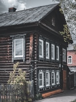 A rustic wooden house with log walls and white-framed windows decorated with intricate carvings. The roof is slanted and made of corrugated metal, and there is a small plant-covered wooden fence in the foreground. A cloudy sky and a portion of a red brick building are visible in the background.