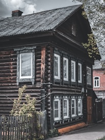 A rustic wooden house with log walls and white-framed windows decorated with intricate carvings. The roof is slanted and made of corrugated metal, and there is a small plant-covered wooden fence in the foreground. A cloudy sky and a portion of a red brick building are visible in the background.