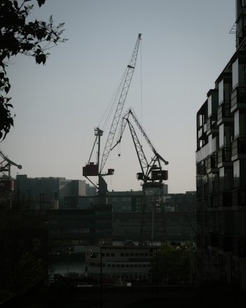 Several large construction cranes are seen against the backdrop of an industrial area with buildings. There is a body of water in the foreground with boats docked. On the right side, there is a tall building with glass windows and balconies, while foliage frames the image on the left side.