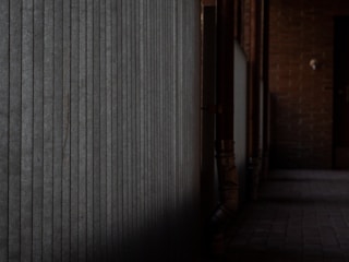 An industrial corridor with a textured metal wall on the left and brick walls on the right. The floor is paved with tiles, and pipes run along the walls. The lighting is dim, casting shadows and creating a moody atmosphere.