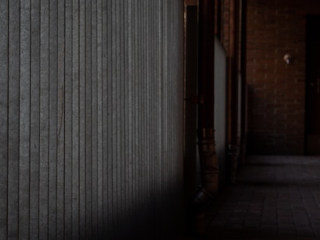 An industrial corridor with a textured metal wall on the left and brick walls on the right. The floor is paved with tiles, and pipes run along the walls. The lighting is dim, casting shadows and creating a moody atmosphere.