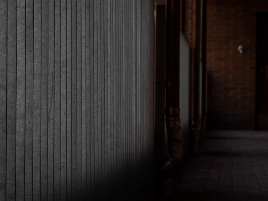 A dimly lit industrial corridor slick with rain and oil, pipes and wires twisting into darkness under flickering fluorescent lights.