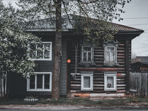 A rustic wooden house with ornate window frames, surrounded by trees. The building features a combination of horizontal log construction and vertical siding. The foliage partially covers the structure, and a small number sign is visible next to the entrance.