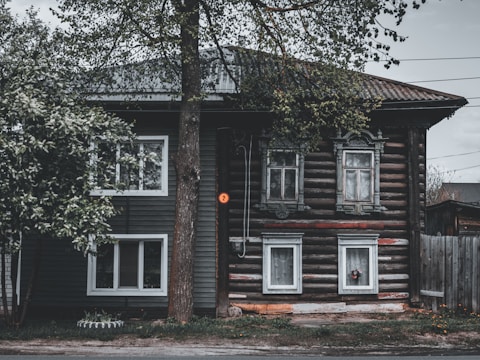 A rustic wooden house with ornate window frames, surrounded by trees. The building features a combination of horizontal log construction and vertical siding. The foliage partially covers the structure, and a small number sign is visible next to the entrance.