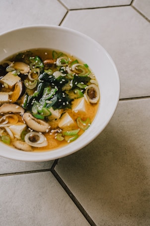 A bowl of miso soup features sliced mushrooms, tofu cubes, seaweed, and sliced green onions, garnished with sesame seeds. The soup is placed on a tiled surface.