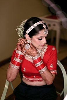A woman in traditional attire with intricate henna designs on her hands and arms. She wears a red blouse adorned with embroidery, complemented by a set of red and gold bangles. Her hair is styled elegantly with a decorative headpiece and flowers. Her earrings and necklace add to the overall embellished look.