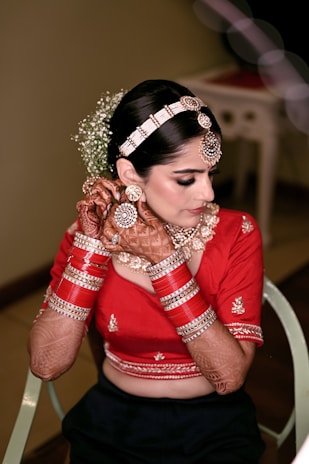 A woman in traditional attire with intricate henna designs on her hands and arms. She wears a red blouse adorned with embroidery, complemented by a set of red and gold bangles. Her hair is styled elegantly with a decorative headpiece and flowers. Her earrings and necklace add to the overall embellished look.