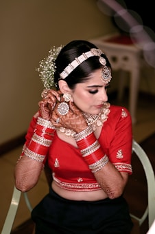 A woman in traditional attire with intricate henna designs on her hands and arms. She wears a red blouse adorned with embroidery, complemented by a set of red and gold bangles. Her hair is styled elegantly with a decorative headpiece and flowers. Her earrings and necklace add to the overall embellished look.