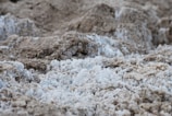 A textured landscape consisting of rough, uneven surfaces in shades of light and dark beige, interspersed with patches of white. The granular appearance suggests a natural, earthy material, perhaps resembling dry sand or salt deposits.