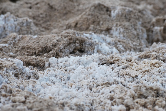 A textured landscape consisting of rough, uneven surfaces in shades of light and dark beige, interspersed with patches of white. The granular appearance suggests a natural, earthy material, perhaps resembling dry sand or salt deposits.