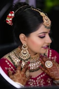 A woman is adorned in traditional bridal attire, featuring intricate jewelry such as a heavy necklace, large earrings, and a forehead ornament known as a maang tikka. Her hair is styled in an updo with decorative flowers. Her hands are beautifully decorated with henna designs. The makeup is elaborate, highlighting her eyes and lips.