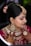A woman is adorned in traditional bridal attire, featuring intricate jewelry such as a heavy necklace, large earrings, and a forehead ornament known as a maang tikka. Her hair is styled in an updo with decorative flowers. Her hands are beautifully decorated with henna designs. The makeup is elaborate, highlighting her eyes and lips.