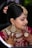 A woman is adorned in traditional bridal attire, featuring intricate jewelry such as a heavy necklace, large earrings, and a forehead ornament known as a maang tikka. Her hair is styled in an updo with decorative flowers. Her hands are beautifully decorated with henna designs. The makeup is elaborate, highlighting her eyes and lips.