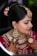 A woman is adorned in traditional bridal attire, featuring intricate jewelry such as a heavy necklace, large earrings, and a forehead ornament known as a maang tikka. Her hair is styled in an updo with decorative flowers. Her hands are beautifully decorated with henna designs. The makeup is elaborate, highlighting her eyes and lips.