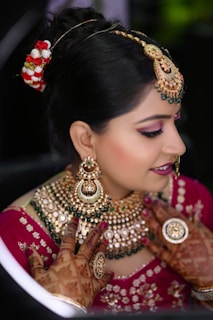A woman is adorned in traditional bridal attire, featuring intricate jewelry such as a heavy necklace, large earrings, and a forehead ornament known as a maang tikka. Her hair is styled in an updo with decorative flowers. Her hands are beautifully decorated with henna designs. The makeup is elaborate, highlighting her eyes and lips.