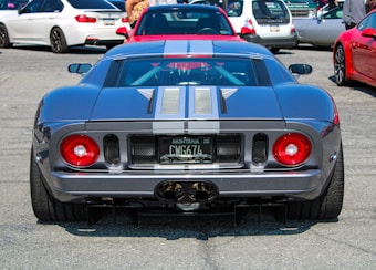 A sports car with distinctive bright red circular tail lights and dual exhaust pipes is parked among other vehicles. The car has a sleek profile and a dark metallic paint job with white racing stripes.