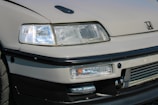 A detailed view of the front end of a car featuring a prominent headlight design, sleek lines, and a recognizable logo. The bumper and grill area are visible, showcasing a sporty aesthetic with a hint of modern design.