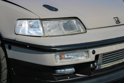 A detailed view of the front end of a car featuring a prominent headlight design, sleek lines, and a recognizable logo. The bumper and grill area are visible, showcasing a sporty aesthetic with a hint of modern design.