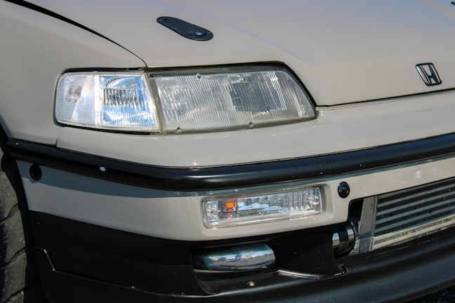A detailed view of the front end of a car featuring a prominent headlight design, sleek lines, and a recognizable logo. The bumper and grill area are visible, showcasing a sporty aesthetic with a hint of modern design.