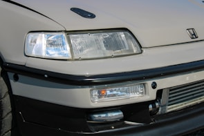 A detailed view of the front end of a car featuring a prominent headlight design, sleek lines, and a recognizable logo. The bumper and grill area are visible, showcasing a sporty aesthetic with a hint of modern design.